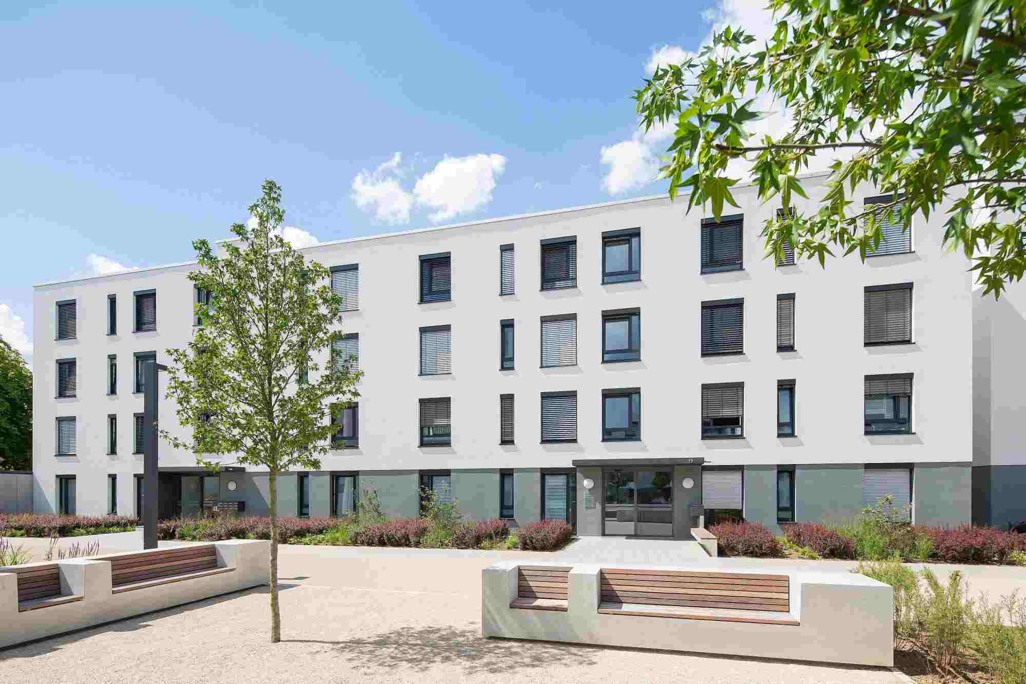 Wohngebäude des Cavalier Holstein in Mainz, weiße Fassade und Flachdach, Sitzbänke und Bäume im Vordergrund